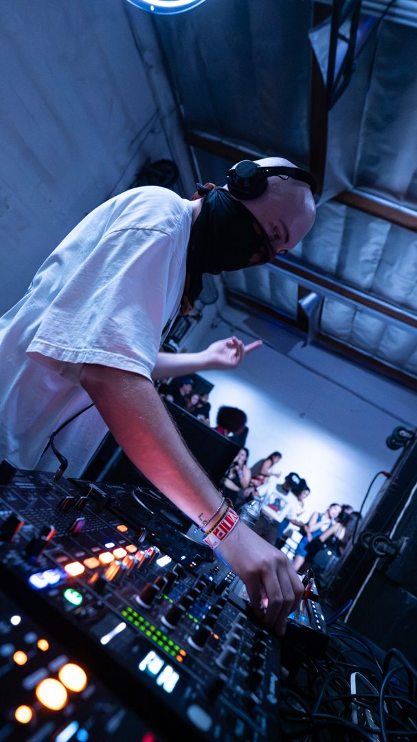 NOWHERE INC., face covered by a black mask, leaning over the mixer mid-set